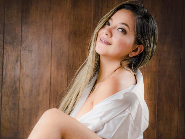 AbrilClark, com cabelos longos loiros e castanhos, camisa branca com um ombro descoberto, mostrando os seios, sorrindo para a câmera, fundo de madeira..
