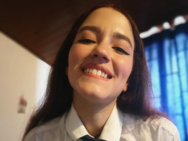 RukkiaParist with long brown hair, smiling, wearing a white shirt with a black tie, has visible white teeth, and slightly flushed cheeks, in a low-angle shot.