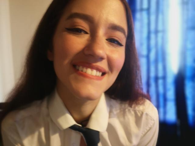 RukkiaParist, met rood haar, lachend, draagt een witte blouse en een zwarte stropdas, heeft eyeliner en rode lippenstift, en ziet er schattig uit met haar tanden zichtbaar. Vage blauwe achtergrond..