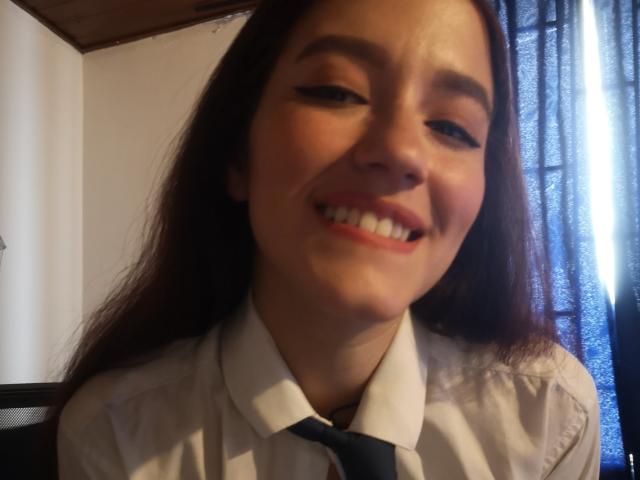 RukkiaParist with long brown hair, smiling with white teeth, wearing a white shirt and black tie, has light makeup and is slightly squinting in the photograph.