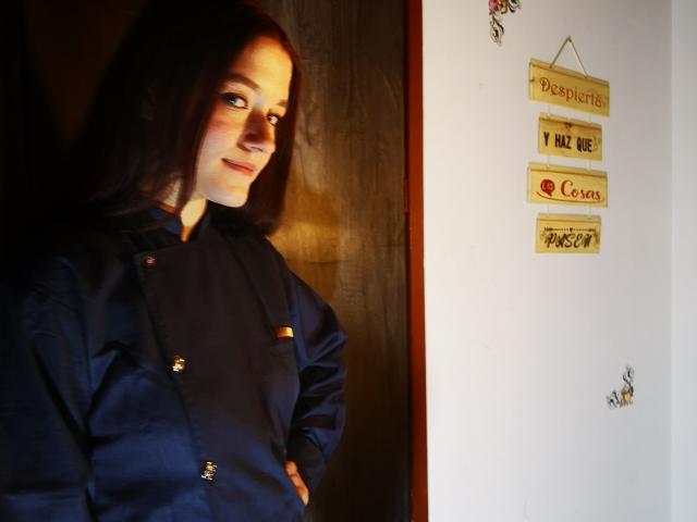RukkiaParist with dark brown hair, wearing a navy blue button-up shirt, hands on hips, slight smile, looking to the side. Wooden wall on left.