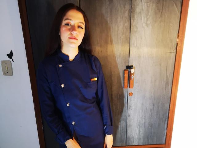 RukkiaParist, with long brown hair, wearing a blue double-buttoned nurse uniform, stands against a wooden door, looking slightly to the side with a neutral expression.