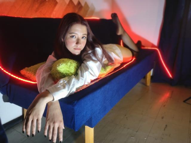 RukkiaParist, recostada en un sofá azul con luces LED rojas, vestida con una camisa blanca, calcetines negros y sosteniendo almohadas verdes, tiene el cabello largo y castaño y esmalte de uñas negro..