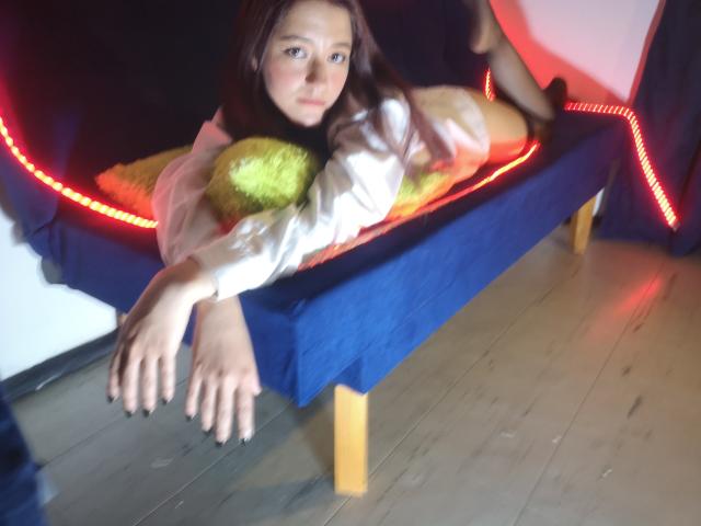 RukkiaParist recostada en un sofá azul con iluminación LED roja, vestida con una camisa blanca, sosteniendo un cojín amarillo, con las manos en el borde del sofá, suelo de madera..