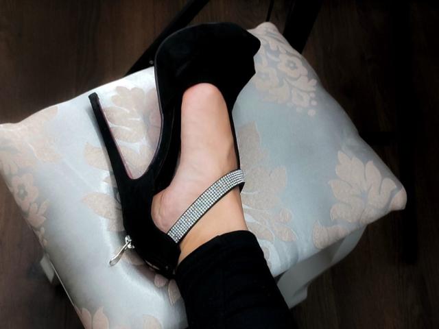 MistressMica's Fuß in einem schwarzen High Heel-Schuh mit einem glitzernden Riemen am Knöchel, ihr schwarzer Hosenbeins teilweise sichtbar, ruht auf einem mit Blumenmuster versehenen Kissen auf einem dunklen Holzboden..