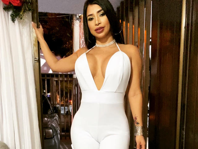 FransheskaParker with large boobs, deep cleavage in a white, tight, sleeveless dress, black hair, choker, standing, hand on door, wooden wall, evening, confident, sexy.
