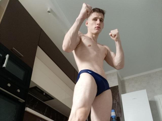 PoligloJoe, standing in a kitchen, wearing blue boyshorts, flexing muscular arms, looking serious, with a slim, toned body, short brown hair, and pale skin.