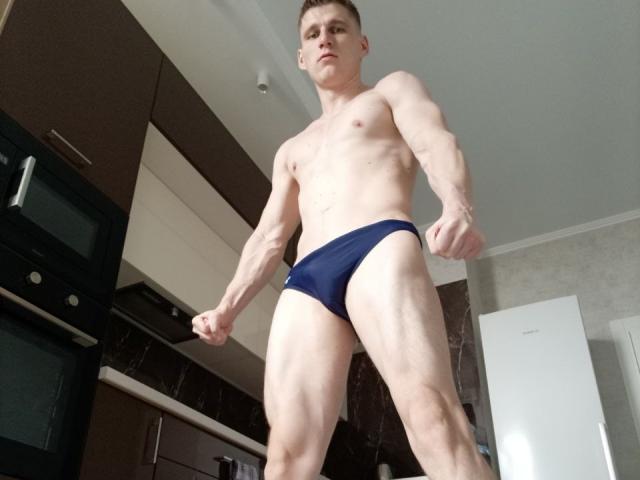 Muscular PoligloJoe with pale skin, standing in a kitchen, wearing tight blue underwear, fists clenched, looking down, short brown hair, visible abs, and defined legs.