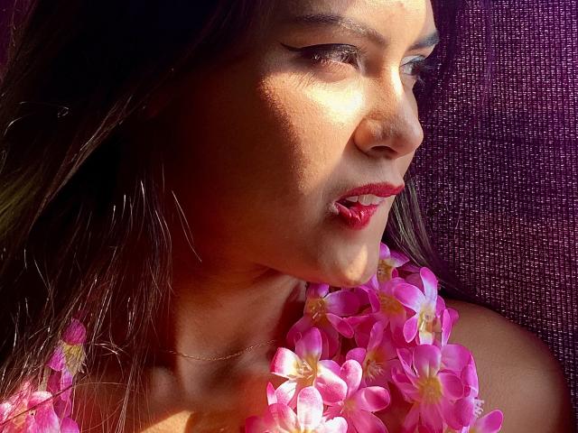 Annies Gesicht, im Sonnenlicht, mit rosa Blumen um ihren Hals, dunklem Haar, braunen Augen, rotem Lippenstift und einer goldenen Halskette, blickt zur Seite. Ihr Hintern ist teilweise sichtbar..