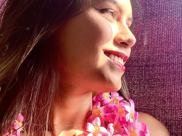 AnnieHal, com cabelo castanho, sorrindo, usando um colar de flores rosa, com luz do sol no rosto, batom rosa, dentes visíveis, uma leve sombra na bochecha direita, fundo roxo texturizado..