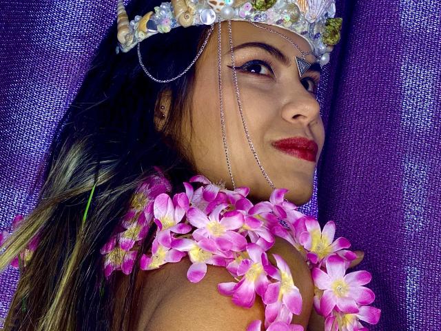 AnnieHal mit rosa Blumen am Hals, die einen mit Juwelen besetzten Kopfschmuck, eine Kette und roten Lippenstift trägt, lächelt und hat dunkles Haar und ein violettes Kleidungsstück. Ihre Brüste sind nicht sichtbar..