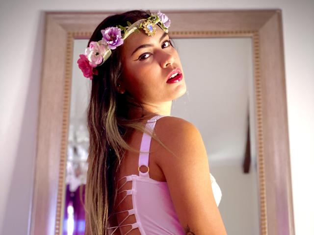 AnnieHal, con lunghi capelli castani, un copricapo a forma di fiore rosa e un abito bianco senza schienale, che mostra il seno, guarda oltre la spalla con il rossetto rosso, in piedi di fronte a uno specchio..