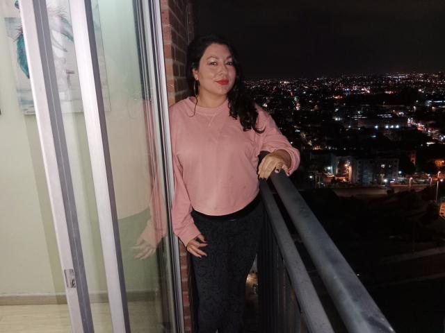 SakotoDika with dark curly hair, pink shirt, and black pants, standing on a balcony at night, leaning on the railing, hands on hips, city lights in the background.