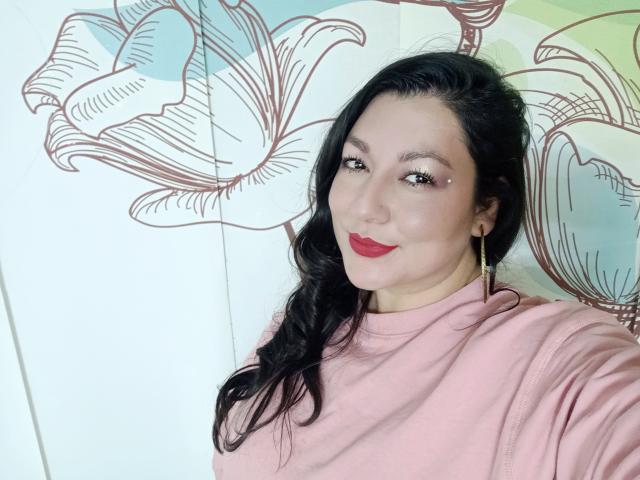 SakotoDika, con i capelli neri, il rossetto rosso, gli orecchini dorati e un maglione a collo alto rosa, sorride alla fotocamera, mostrando un piccolo piercing sul naso sul lato sinistro..