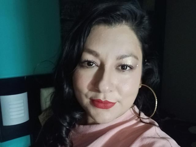SakotoDika with shiny eyes, red lipstick, and a pink turtleneck, wearing gold hoop earrings, has black wavy hair, and a slight smile, looking at the camera.