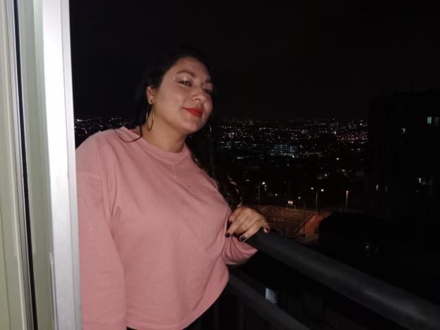 SakotoDika, com cabelo escuro, suéter rosa, batom vermelho, um pequeno piercing no nariz e brincos de argola, apoiada na varanda à noite, com as luzes da cidade ao fundo, a mão na grade, com um leve sorriso..