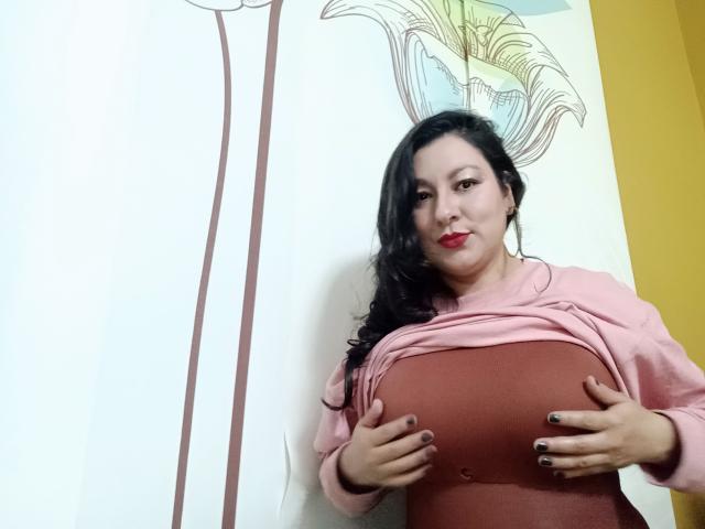 SakotoDika with dark hair, red lipstick, pink sweater, and brown top, holding her large tits with painted nails, standing against a wall with a floral drawing.