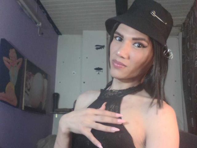 LunaGransTs con una parte superiore nera in pizzo, una mano sui seni, indossa un cappello nero con un anello d'argento, rossetto rosa e eyeliner, in una stanza con pareti viola. Ha la pelle media e i capelli scuri..