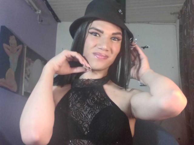 LunaGransTs, with black lace halter top, showing off her tits, wearing a black hat, smiling, hands near ears, long black hair, light skin, and subtle makeup, in a dim room.