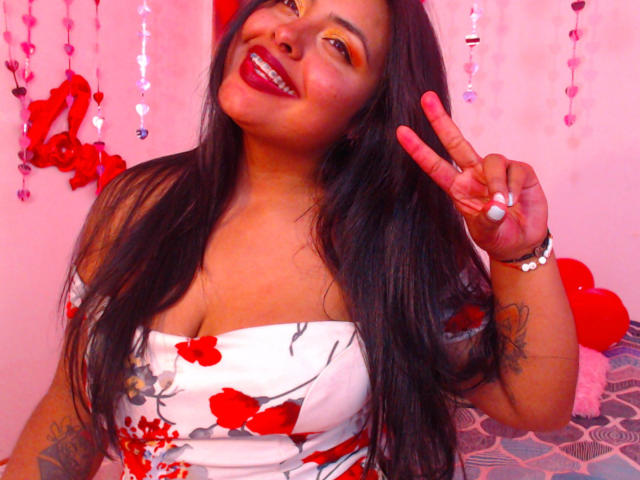 Angela Roggers mit langem, schwarzem Haar, rotem Lippenstift und einem weißen Kleid mit einem Träger, das rote Blumenmuster aufweist, zeigt ein Friedenszeichen, ihre Brüste und lächelt..