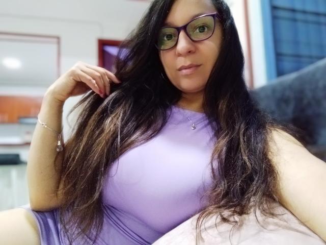 Angelonix with long dark hair, wearing purple glasses and a tight purple top, showing big boobs, sitting and touching her hair with a relaxed smile.