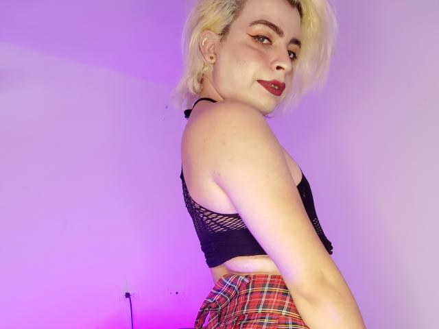 TsFoxCandy con piel pálida, cabello rubio, lápiz labial rojo, top de malla negra y pantalones de cuadros rojos, mirando por encima del hombro, mostrando un poco de su trasero. Iluminación púrpura en el fondo..