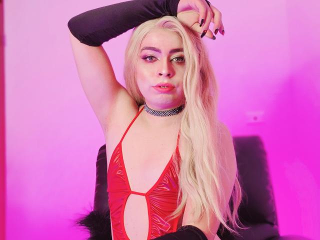 TsFoxCandy with long blonde hair, red vinyl cut-out top, black choker, black arm sleeves, and pink neon lighting, raising her right arm, showing her tits.