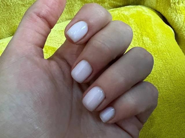 La mano de Innysik, con esmalte blanco brillante en uñas cuidadosamente recortadas, sobre un fondo amarillo texturizado y brillante, mostrando una piel clara y sutiles tonos rosados..
