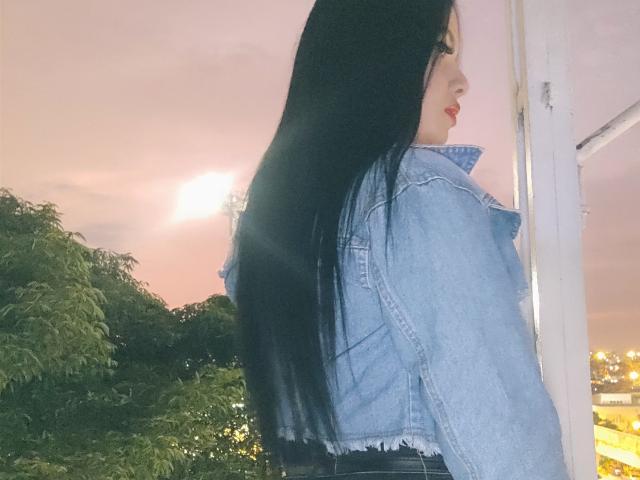 AshleyKatelyn met lang zwart haar, die een blauwe denimjas draagt, kijkt over haar schouder, met zonlicht in haar haar, rode lippenstift, staat in de buurt van een witte deur, groene bomen..