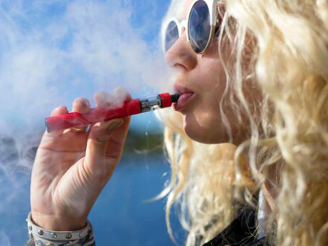 Vikki, com cabelo loiro encaracolado, usando óculos de sol redondos, fumando um cigarro vermelho, céu azul ao fundo, língua ligeiramente para fora, pele clara, jaqueta preta, pulseira de prata no pulso..