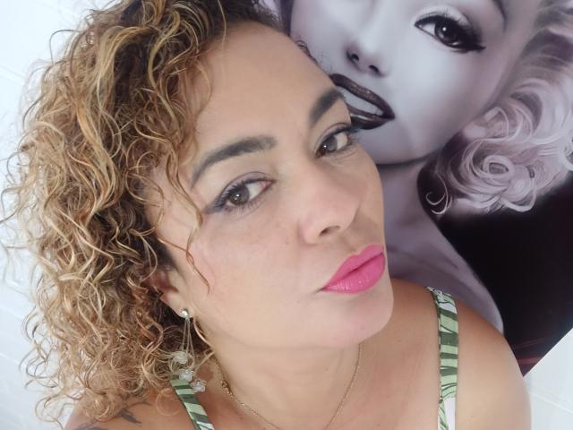 SolmaryHot with curly blonde hair, pink lipstick, green strap top, and hoop earrings, looking down with smoky eye makeup, next to a Marilyn Monroe poster.