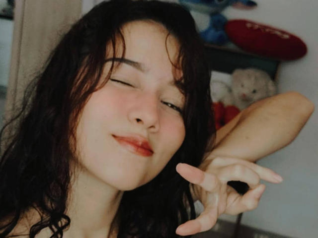 DeisyWest with wavy black hair, eyes closed, smiling, and pointing with her right hand. She has red lipstick and a cute, relaxed expression. Her ass is not visible.