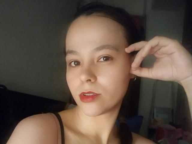 HotJoyLover with smooth, fair skin, dark brown hair in a braid, red lipstick, brown eyes, small nose, black tank top, right hand touching ear, subtle smile, slight shadow.