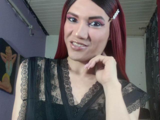 LunaGransTs with red hair, black lace top, pink eyeshadow, glossy lips, smiling, hand near chin, hairclip, pale skin, medium breasts, white ceiling, purple wall.
