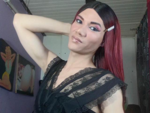 LunaGransTs con capelli rossi intrecciati, vestito di pizzo nero e eyeliner rosa, che fa la smorfia con un braccio dietro la testa, labbra lucide, pelle pallida, piccoli seni, in una stanza poco illuminata..