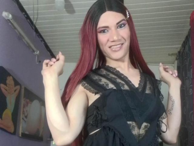 LunaGransTs con cabello rojo, vestido de encaje negro, sonriendo, con las manos levantadas, mostrando tatuajes en su brazo izquierdo, en una habitación con paredes moradas y un ventilador de techo..