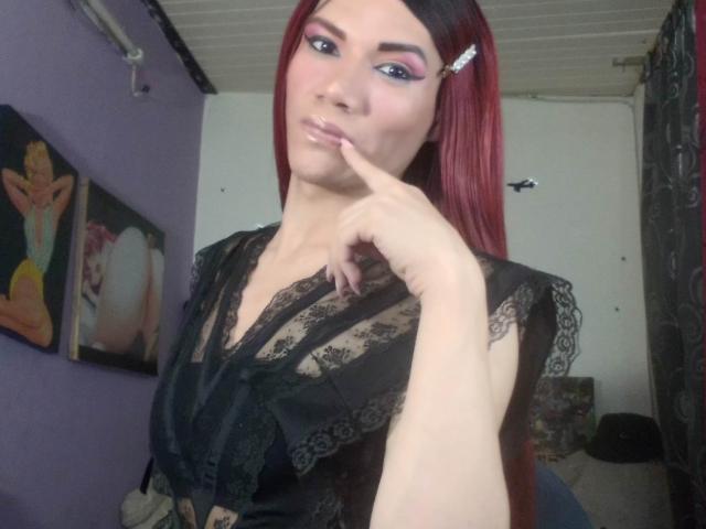 LunaGransTs with red hair, black lace top, finger on lips, pink eyeshadow, white hair clip, light skin, slight smile, in a purple-walled room with art.