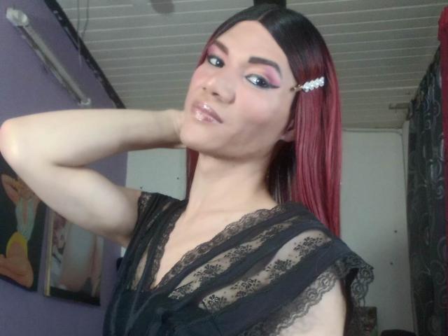LunaGransTs with red and black hair, wearing black lace top, pouty lips, pink eyeshadow, and a hair clip, posing with hand behind head, smiling slightly.