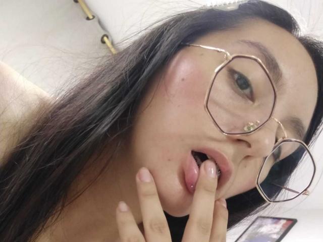 LatinAleja, con cabello negro largo, mejillas rosadas y gafas redondas, chupándose el dedo, con la lengua afuera, con una expresión seductora, piel clara, boca ligeramente abierta, labios rosados suaves y uñas rosas..