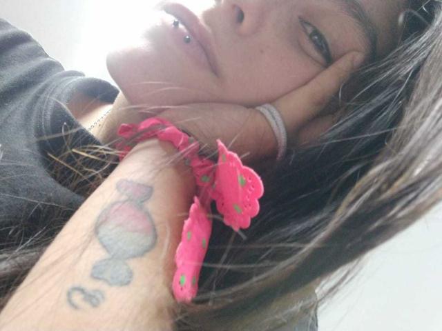 LatinAleja, con un piercing en la nariz, delineador de ojos negro y una goma elástica rosa en la muñeca, tiene un tatuaje en el antebrazo y está apoyando la cabeza en la mano..