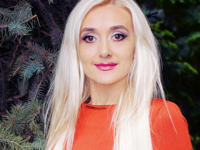 SophieKity ha i capelli biondi, il rossetto rosa e il trucco agli occhi viola. Indossa una maglietta rossa, appare carina con il seno e i glutei leggermente visibili. Sta sorridendo..