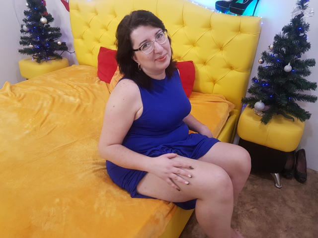 HelenEnigma, seduta su un letto giallo, indossa un vestito blu, occhiali e sorride, con le mani appoggiate sulle cosce, mostrando il sedere. Due piccoli alberi di Natale sono posti su entrambi i lati..