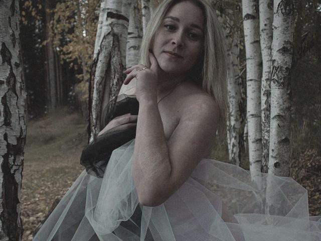 DoraSevi, mit heller Haut, blonden Haaren und kleinen Brüsten, sitzt in einem Wald und trägt ein durchsichtiges, schwarz-weißes Kleid. Sie hält ihre Brust mit einem sanften, verführerischen Lächeln..