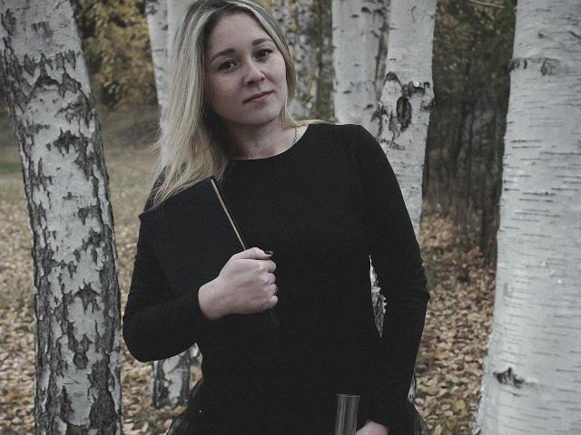 DoraSevi mit blonden Haaren, in einem schwarzen, langärmeligen Kleid, hält einen Stock, steht inmitten von weißen Birken, lächelt leicht, Herbstlaub auf dem Boden..