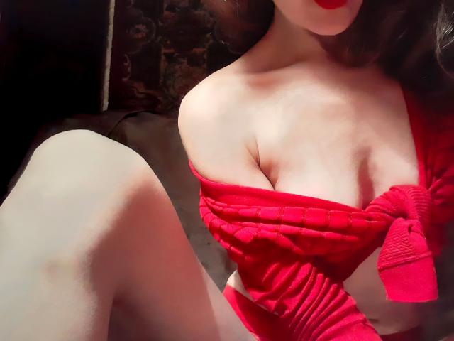 Le pull rouge à épaules dénudées d'Orshiti est tiré vers le bas, exposant ses grandes seins et sa poitrine. Elle porte des sous-vêtements rouges assortis, et une de ses jambes est relevée au premier plan..