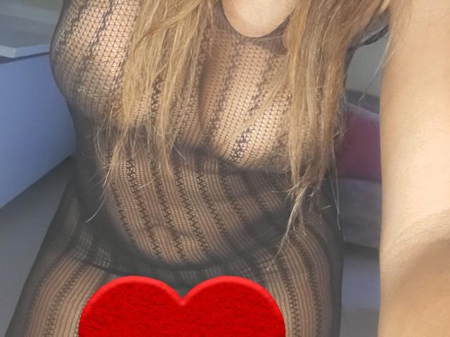 AnaSexy wearing a see-through black lace dress, her tits visible through the fabric, blonde hair, red heart covering her pussy, arm partially visible.
