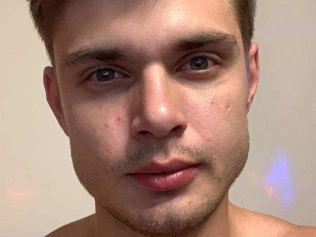 MaksWolfis tem um rosto com algumas espinhas, levemente brilhante, com cabelo castanho curto, olhos verdes e uma barba clara. Seus lábios rosados estão ligeiramente entreabertos. Ele está sem camisa, mostrando seu pescoço nu..