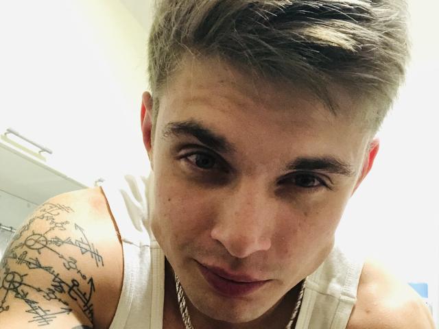 MaksWolfis tiene el pelo corto y canoso, ojos marrones y un tatuaje en el hombro derecho. Lleva una camiseta blanca y una cadena de plata. Está mirando a la cámara..