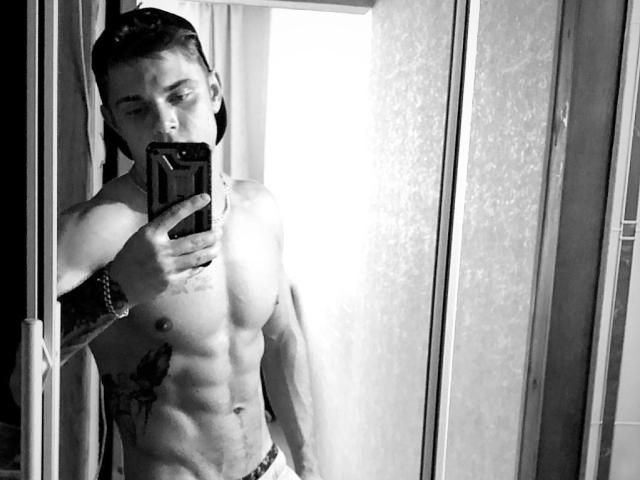 Selfie in bianco e nero di MaksWolfis, con il torso muscoloso e tatuato scoperto, che tiene un telefono, indossa biancheria intima bianca e ha i capelli intrecciati. Gli addominali di MaksWolfis sono visibili..