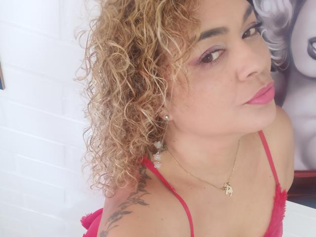 SolmaryHot has curly blonde hair, pink lipstick, red dress, tattoos on her shoulder, and a gold necklace with a small pendant. She's looking up with a slightly pouty face.